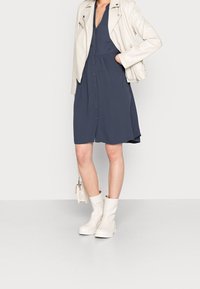 Kvinna i en marinblå klänning med knappar fram, krämfärgad läderjacka i bikerstil, vita ankelskors och håller en liten krämfärgad handväska.