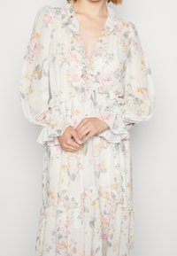 Femme en robe blanche à fleurs à manches longues avec un col et des poignets à volants, les mains délicatement croisées à la taille.