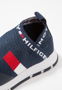 Zapatilla navy de slip-on con textura de punto, que presenta detalles en blanco, rojo y azul. Suela blanca con detalles de tracción en negro. Parte superior de tela suave.