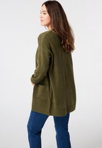 Olijfgroene, oversized gebreide cardigan met lange mouwen, geribbelde zoom en voorzakken. Draag met blauwe denimbroek.