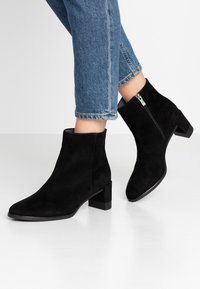 Bottines en daim noires avec un bout carré, un petit talon carré et une fermeture éclair sur le côté. Associées à un jean en denim bleu coupé.