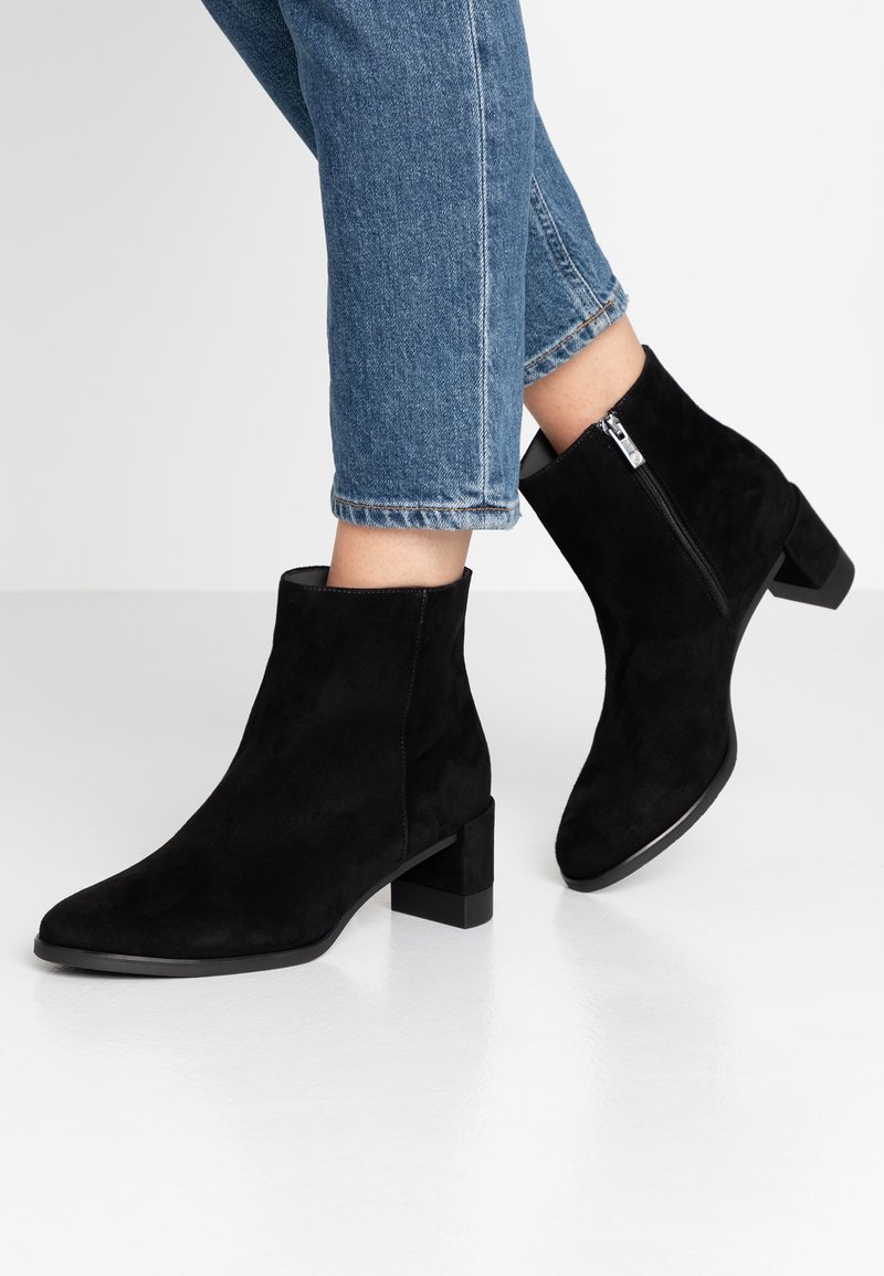 Bottines en daim noires avec un bout carré, un petit talon carré et une fermeture éclair sur le côté. Associées à un jean en denim bleu coupé.
