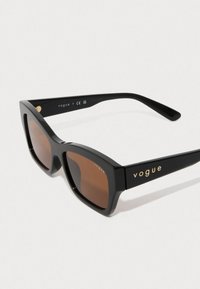Čierne slnečné okuliare s obdĺžnikovým tvarom, hnedými sklami a zlatým brandingom "vogue" na stranách. Hladké plastové rámiky s rovnými hranami.
