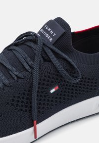 Marineblau gestrickter Sneaker mit strukturiertem Obermaterial, runden Schnürsenkeln, gebrandeter Zunge, weißem Mittelsohl und roten sowie weißen Akzentstreifen an der Seite.