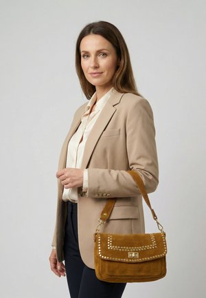 Vrouw die een beige blazer en een witte blouse draagt, met een bruine suède handtas met gouden studs en een schouderriem.