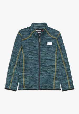 Blaugrüner und schwarzer melierter Reißverschluss-Pullover mit gelben Nahtakzenten und einem kleinen rechteckigen Patch mit schwarzen Punkten auf der linken Brust.