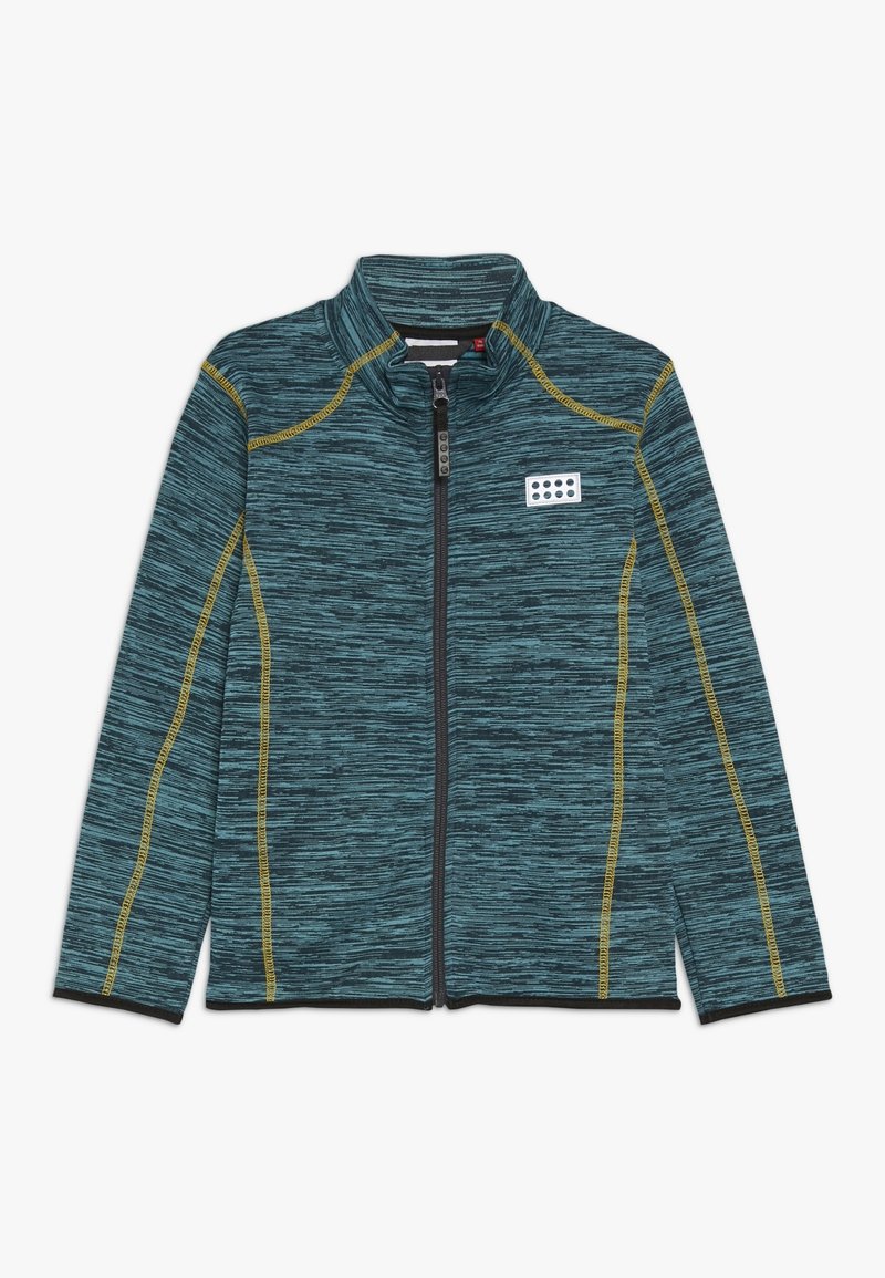 Chaqueta de cremallera en mezcla de azul verdoso y negro, con detalles de costuras amarillas y un pequeño parche rectangular con puntos negros en el lado izquierdo del pecho.