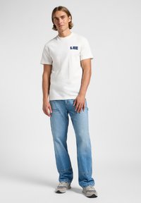 T-shirt bianca in cotone a maniche corte con il logo navy "LEE" sul petto, abbinata a jeans in denim azzurro chiaro e sneakers grigie.
