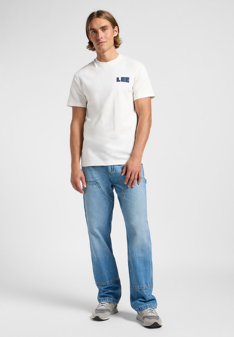T-shirt bianca in cotone a maniche corte con il logo navy "LEE" sul petto, abbinata a jeans in denim azzurro chiaro e sneakers grigie.