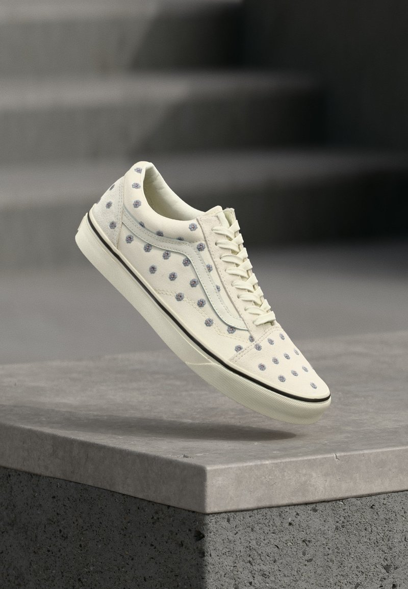 Baskets blanc cassé avec des motifs floraux bleus et rouges, des lacets blancs et une semelle à rayures noires flottant au-dessus d'un bloc de béton gris.