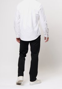 Valkoinen nappikauluspaita, jossa on kaareva helma, yhdistettynä mustiin housuihin ja valkoisiin lenkkareihin. Paidassa on pitkät hihat ja nappirannekkeet.