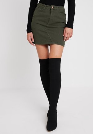 Modèle féminin portant une mini-jupe en denim vert olive, un haut noir à manches longues et des bottes hautes en noir jusqu'aux genoux, se tenant devant un fond uni.