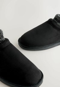 Sorte slippers med en blød ruskindsudvendig, rund tå og en struktureret grå strikket ankelmanschette. Flade såler til indendørs brug.
