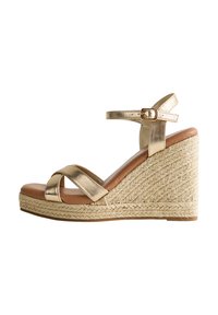 Sandale compensée en métal doré avec des sangles entrecroisées et une boucle à la cheville. Texture en jute tissé sur la compensée, semelle en cuir tan et finition lisse.