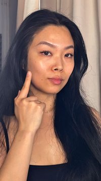 En person med långt svart hår och ljus hy pekar på sin kind. De har subtila fräknar, naturlig sminkning och en enkel silverhalsband.