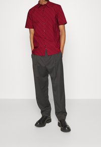 Camisa roja de manga corta con un patrón diagonal, combinada con pantalones grises de corte holgado y zapatos negros con cordones.