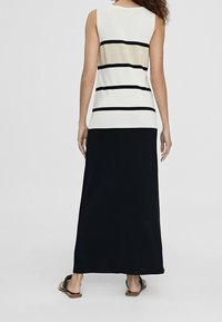 Donna che indossa un abito maxi senza maniche bianco e nero con strisce nere orizzontali e dettagli dorati, abbinato a sandali neri.