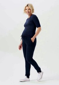 Marineblaue Umstandbluse mit kurzen Ärmeln und Bindedetail, kombiniert mit marineblauen Jogginghosen mit Taschen, getragen mit weißen Sneakers. Sanfte Textur.