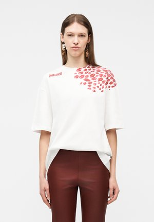 Nő fehér, túlméretezett ingben, amelyen piros ajkak nyomata és a "Just Cavalli" felirat látható a vállán, sötétvörös szűk nadrággal párosítva.