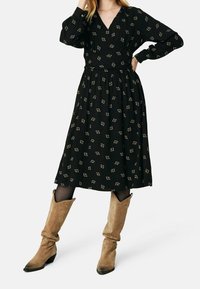 Robe noire à manches longues avec un décolleté en V, ornée de petits carrés dorés ; taille cintrée et jupe fluide, associée à des bottes en daim beige.