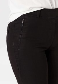 Gros plan d'un pantalon noir avec une poche latérale zippée porté avec un haut blanc, montrant la taille et les détails de couture.