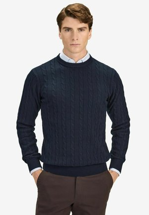 Mørkeblå sweater med et kabelstrikdesign, ribstrikket halsudskæring, manchetter og kant. Bæres over en lyseblå skjorte med krave, der fremviser tekstureret stof.