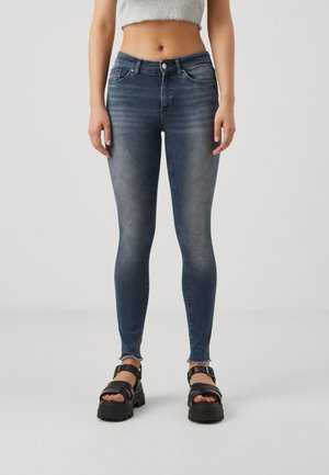 Jeans en denim skinny bleu foncé avec des détails délavés, une légère usure et des ourlets effilochés. Associés à des sandales compensées noires.