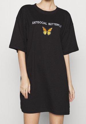 Person trägt ein schwarzes übergroßes T-Shirt-Kleid mit dem Schriftzug "ANTISOCIAL BUTTERFLY" und einem orange-gelben Schmetterling, der auf der Brust eingestickt ist.