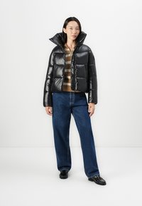 Zwarte pufferjack met glanzende afwerking, cropped ontwerp, verhoogde kraag en ritssluiting aan de voorkant. Gepaarde met gestreepte trui en wijd uitlopende jeans.