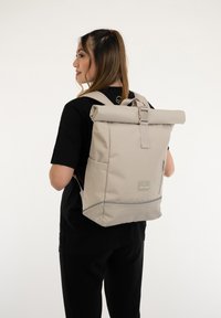 Beige színű, felhajtós hátizsák, állítható vállpántokkal, elülső cipzáras zsebbel és logóval ellátott folttal. Tartós anyagból készült, sima felülettel.