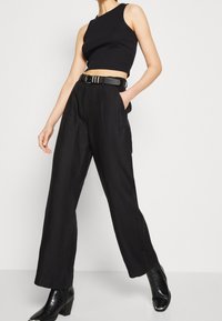 Černý zkrácený top bez rukávů v kombinaci s černými kalhotami s vysokým pasem a širokými nohavicemi. Kalhoty mají pásek s texturovaným kováním. Lesklé černé boty.