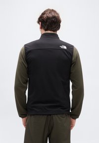 Άνδρας που φοράει μαύρο γιλέκο North Face πάνω από ελιά πράσινο μακρυμάνικο πουκάμισο, σε λήψη από πίσω με φόντο ένα απλό φωτεινό φόντο.