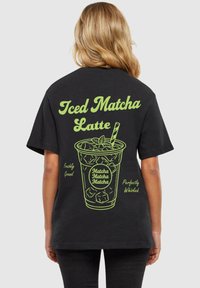Juodas marškinėliai su žalia grafika, vaizduojančia ledinį matcha latte, su tekstu "Ledinė Matcha Latte", "Šviežiai Sumalti" ir "Idealiai Išplakti".