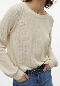 Femme portant un pull en tricot crème avec de subtils motifs verticaux, un jean bleu et une ceinture noire, les mains dans les poches du jean.