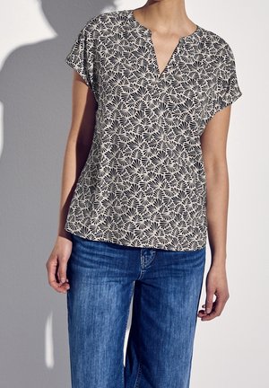 Femme portant un chemisier beige à manches courtes avec un motif en forme d'éventail noir et un jean en denim bleu, debout contre un mur gris clair.