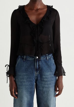 Frau trägt eine transparente schwarze Bluse mit Rüschen und Bändern, kombiniert mit hoch taillierten blauen Jeans vor einem schlichten weißen Hintergrund.