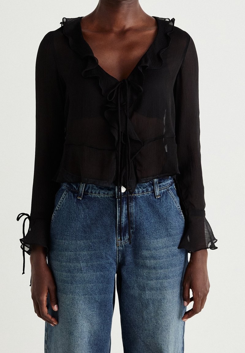 Femme portant un chemisier noir transparent avec des volants et des liens, associé à un jean bleu taille haute sur un fond blanc uni.