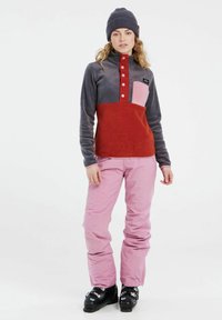 Fleece pullover in donkergrijs en rood met een roze borstzak, met een hoge kraag en knoopsluiting. Gepaard met lichtroze broek en zwarte laarzen.