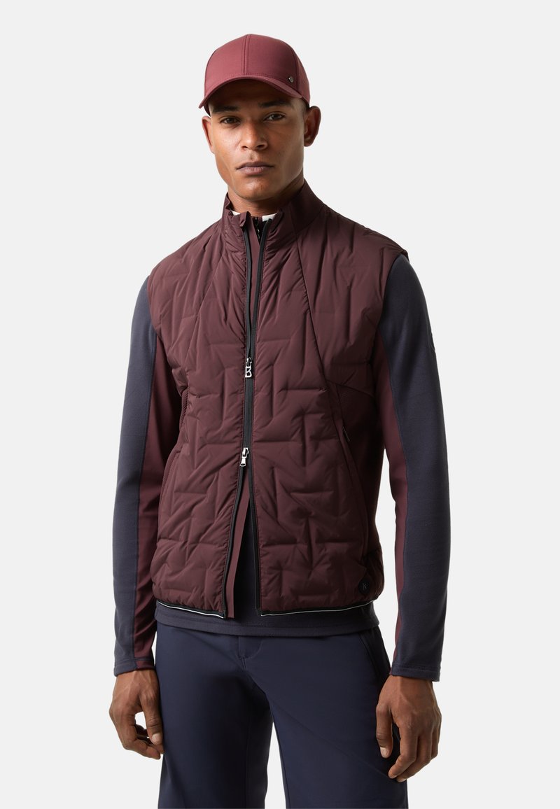 Bordeauxfarbene, wattierte Weste mit Steppmuster, kombiniert mit einem marineblauen Langarmshirt. Ein passendes bordeauxfarbenes Baseballcap rundet das Outfit ab.