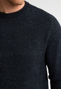 Gebreide trui in donker marineblauw met een gespikkeld patroon. Heeft een geribbelde ronde hals en een textuurstof. Luxe uitstraling met subtiele glans.