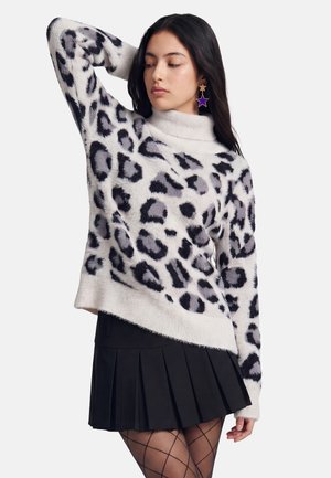 Ung kvinde med langt mørkt hår iført en hvid og grå leopardprint sweater, sort plisseret nederdel, stjerneformede øreringe og mønstrede strømpebukser.