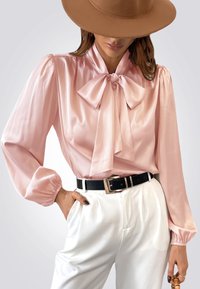 Blusa de satén rosa claro con lazo, mangas abullonadas y textura suave, combinada con pantalones blancos de tiro alto y un cinturón negro.