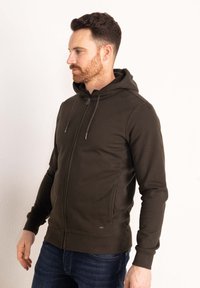 Felpa con zip color verde oliva, dotata di cappuccio regolabile con cordino, maniche lunghe, zip frontale e polsini e orlo a costine.