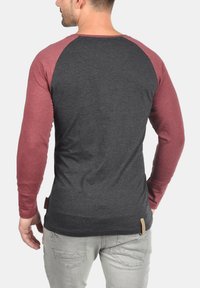 Langarmshirt mit dunkelgrauem Körper und kastanienbraunen Raglanärmeln. Stoff aus Baumwollmischung, mit einem kleinen Markierungslabel am Saum.
