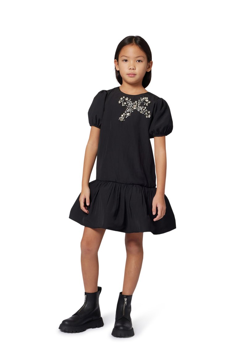 Vestito nero a maniche corte con spalle a sbuffo e gonna a volant. Presenta un'imbottitura di perline sul petto. Indossato con stivali neri alla caviglia.