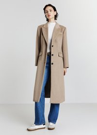 Cappotto lungo beige in misto lana con rever a punta, due tasche frontali e bottoni neri, indossato sopra una maglia bianca e jeans blu svasati.