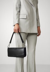 Osoba w jasnoszarym garniturze trzyma czarną torebkę ze skóry wytłaczanej w wzór krokodyla za krótki pasek.