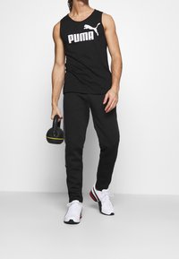 Débardeur noir sans manches avec logo "PUMA" blanc, accompagné d'un pantalon de survêtement noir et de baskets blanches, tenant un kettlebell noir avec un accent jaune.