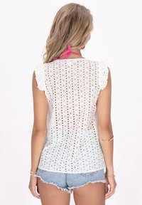 Vit ärmlös topp med broderad design och volangaxlar. Baksidan har en rosa knytning. Materialet är lätt och genomskinligt.