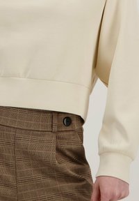 Sudadera color crema con mangas largas y dobladillo acanalado, combinada con pantalones marrones a cuadros que presentan un detalle de botón negro.